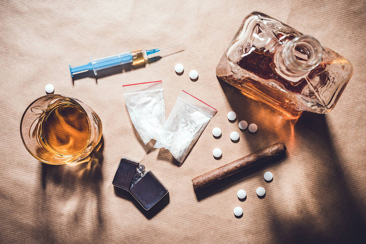Volksdroge Alkohol Eine Spritze, Tabletten, eine Zigarre, ein Glas und eine Flasche Alkohol stehen auf einem Tisch