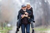 Ein älterer Mann trägt eine Frau Huckepack auf einem Waldweg.