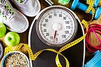 Auf einer Körperwaage liegt ein Maßband, drumherum liegen Hanteln, Sportschuhe und Äpfel