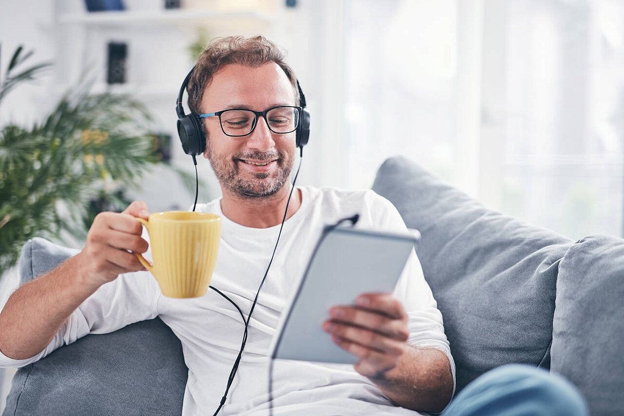 Mann sitzt mit Kopfhörern, Tasse und Tablet auf einem Sofa