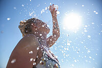 Eine Frau schüttet sich Wasser ins Gesicht. Im Hintergrund blauer Himmel und Sonne.