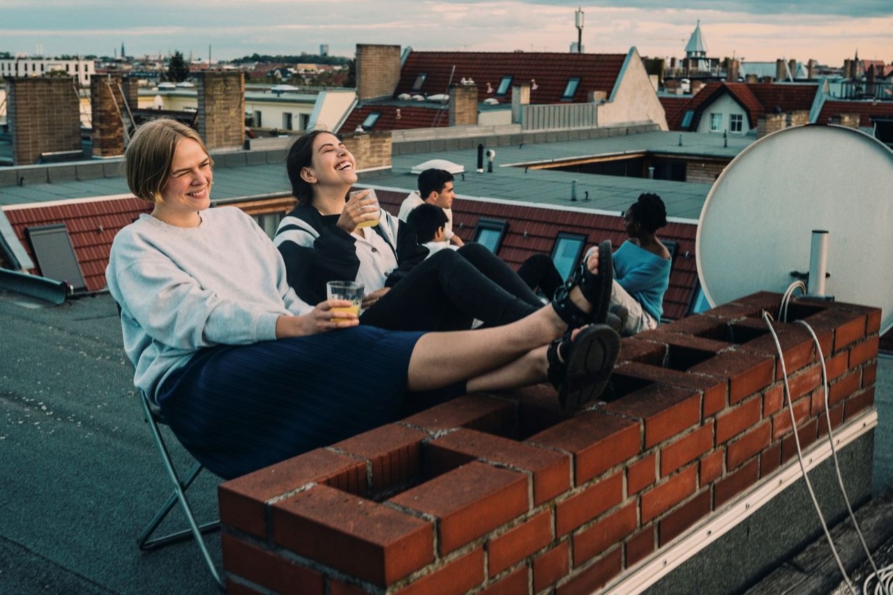 Fünf Jugendliche sitzen lachend auf einem Häuserdach, zwei im Vordergrund trinken ein gelbes Getränk