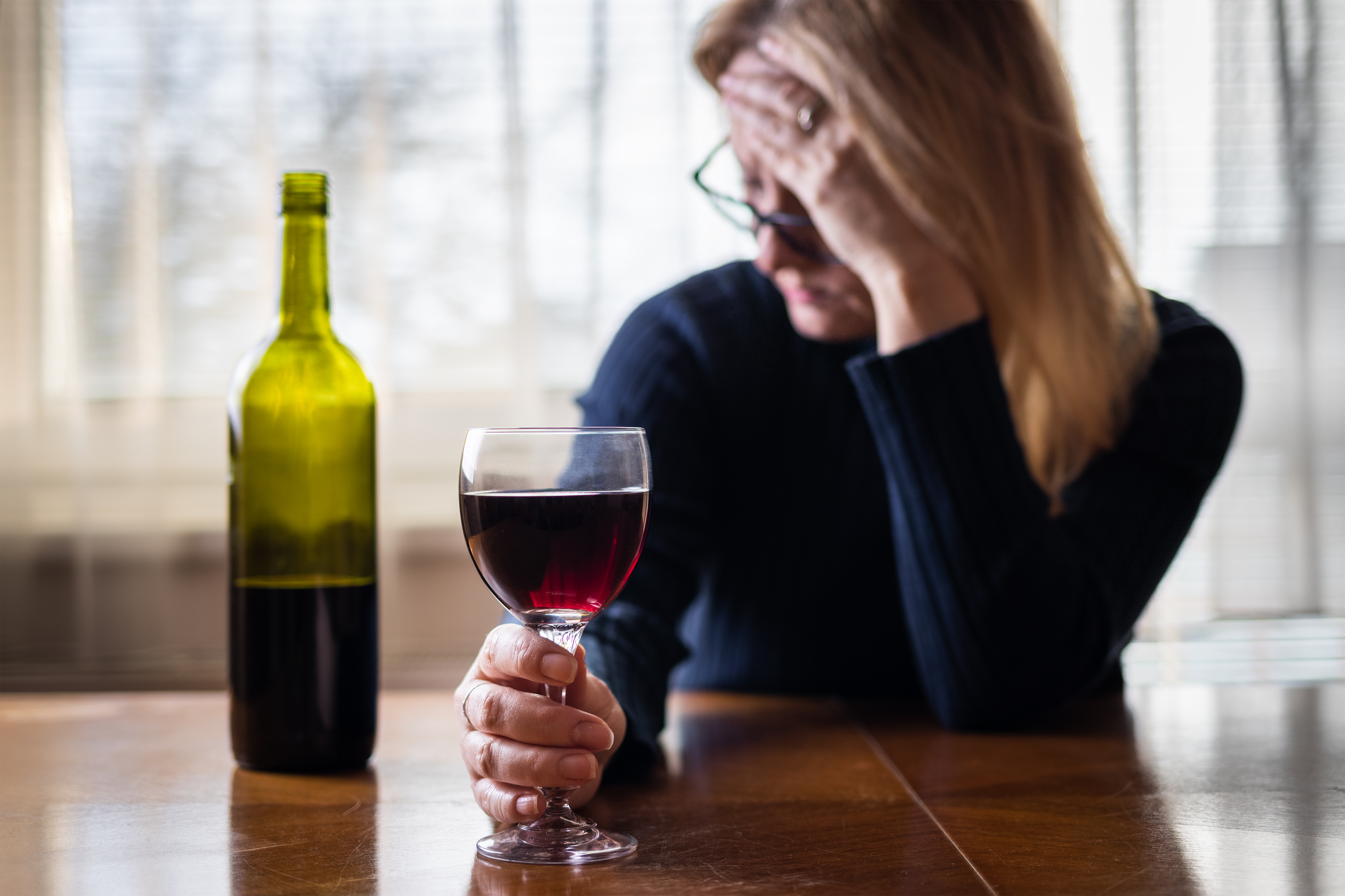 Frau sitzt am Tisch und hält sich mit der einen Hand die Stirn, mit der anderen ein Glas Rotwein