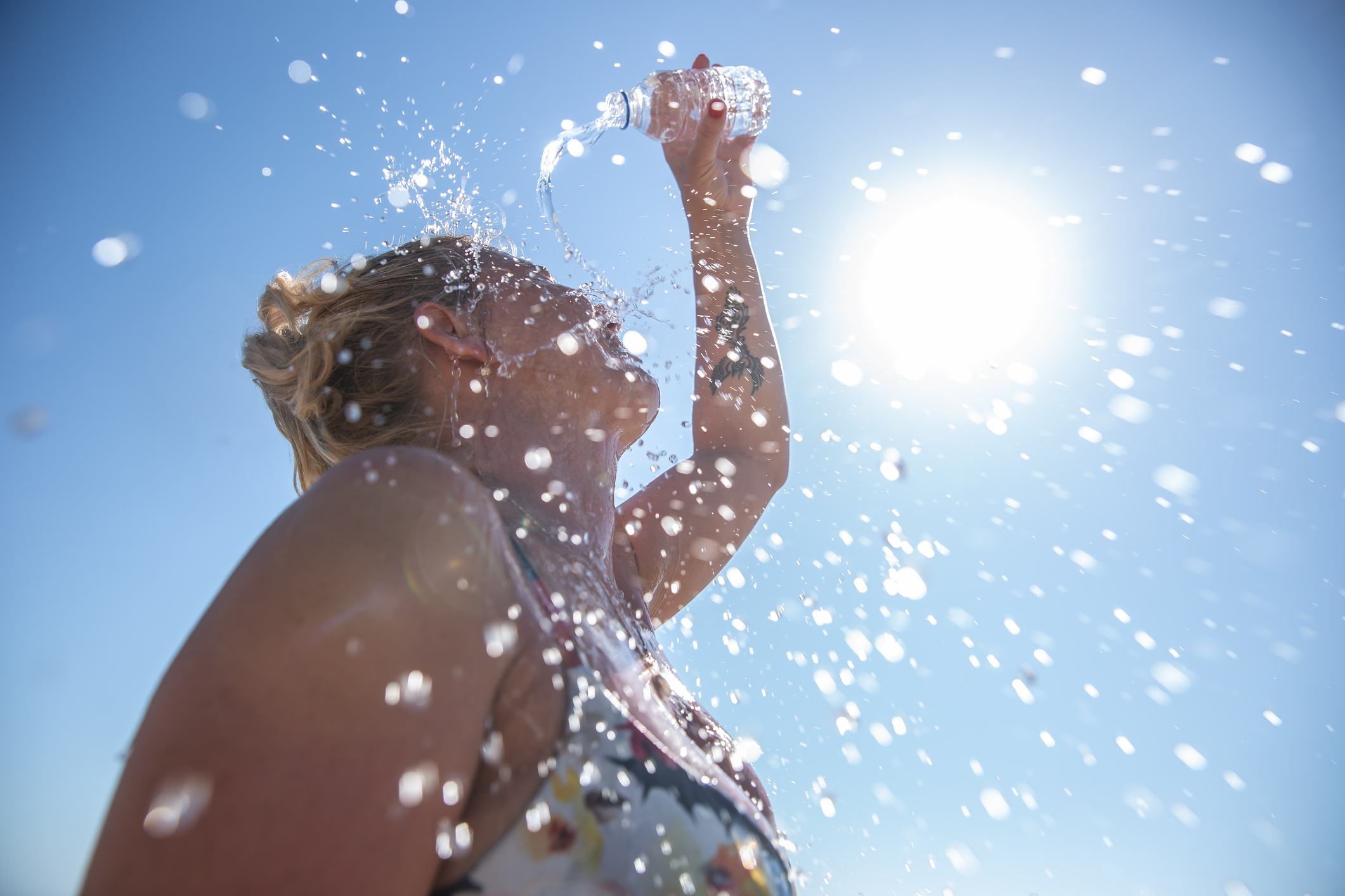 Eine Frau schüttet sich Wasser ins Gesicht. Im Hintergrund blauer Himmel und Sonne.