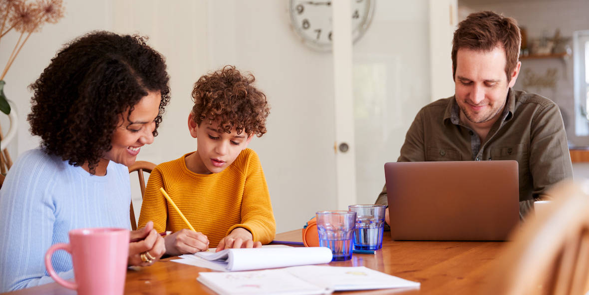 Familie am Tisch: Vater arbeitet am Laptop, Mutter hilft Sohn bei Hausaufgaben